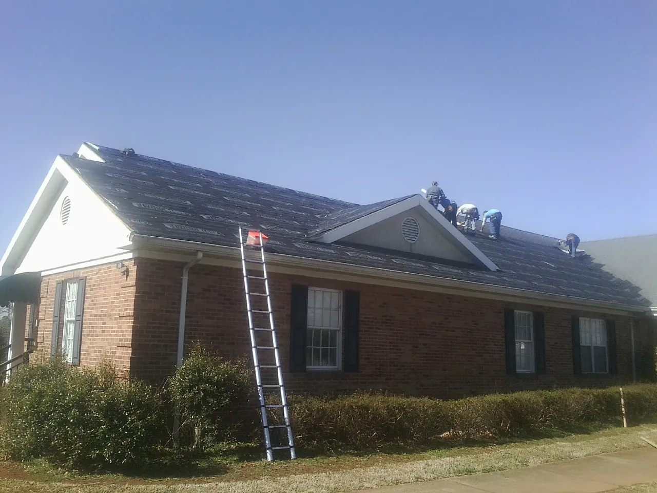 Crew tearing shingles off a brick ranch roof with a ladder set — Emergency Roof Repair work by our crew in Dike