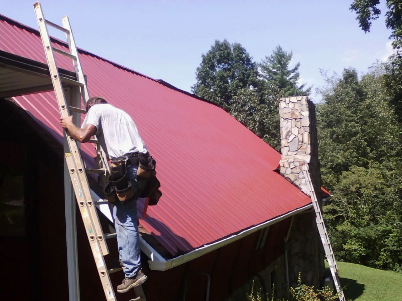Roofer on a ladder beside a red metal roof and stone chimney — Soffit & Fascia Repair by Roofing Brothers in Dike